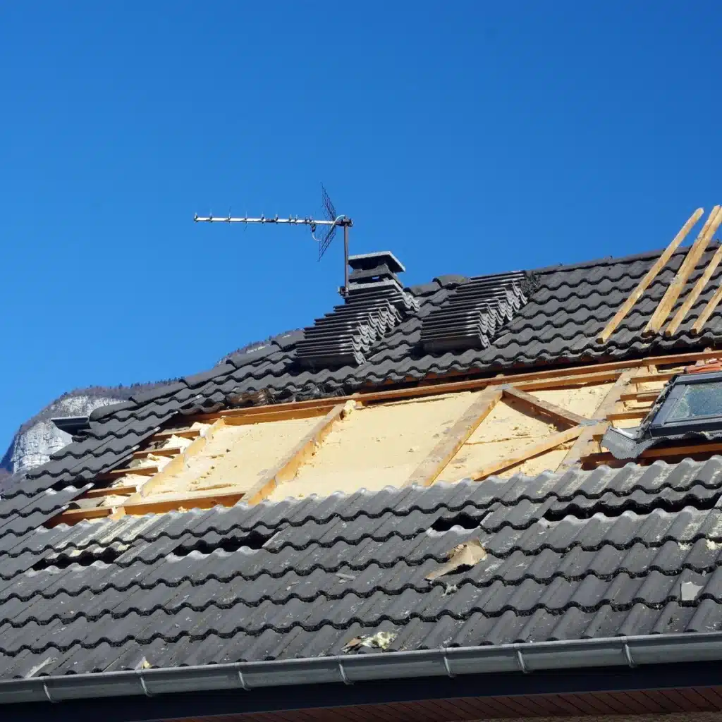 Travaux d'isolation de toiture en cours, avec des tuiles retirées pour accéder à l'isolation et une structure de charpente visible, sous un ciel bleu clair.